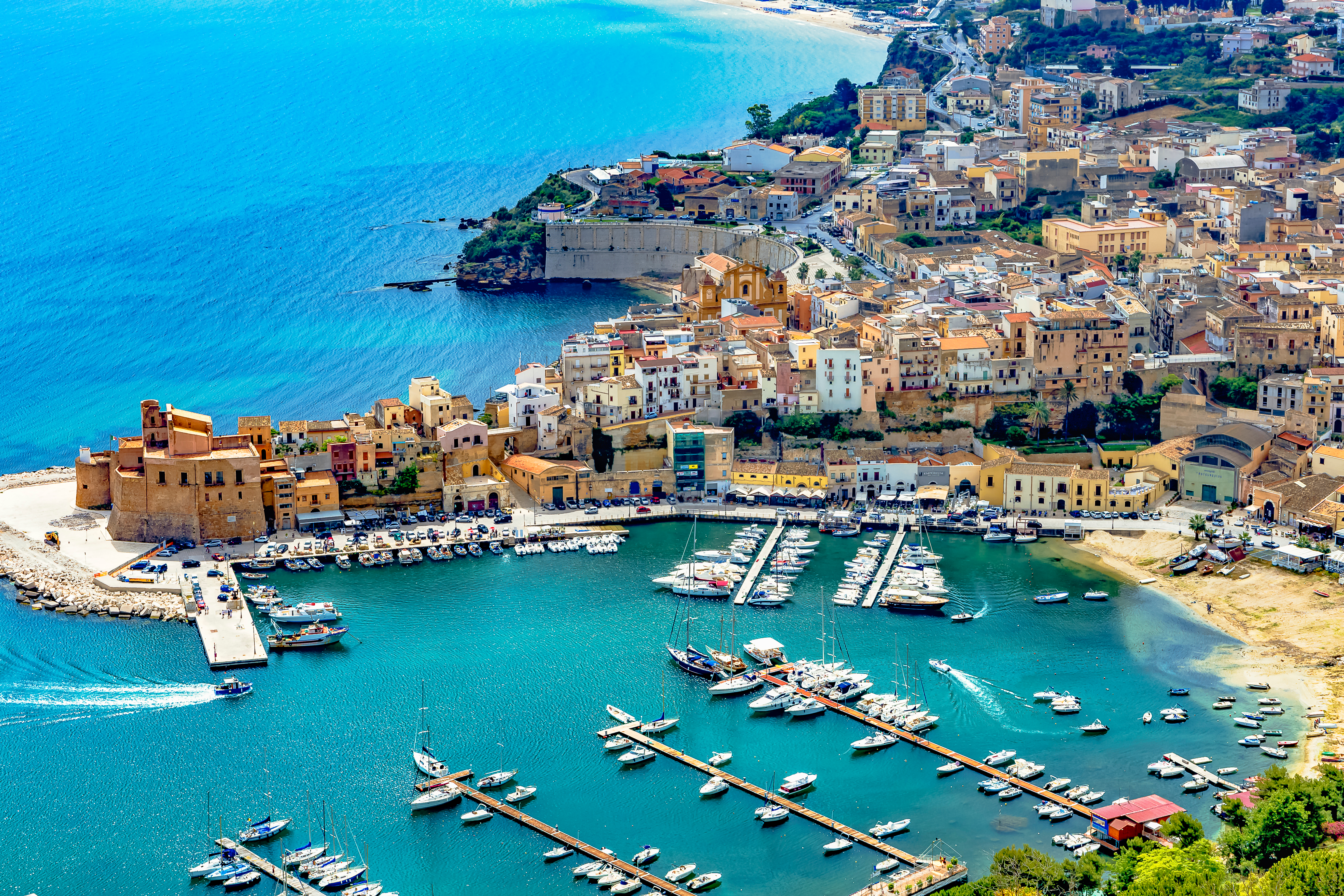 Historic Sicilian port city with colorful buildings lining the waterfront, fishing boats docked along the shore, and distant hills under a bright sky. The scene feels lively and welcoming, evoking a sense of Mediterranean charm and coastal adventure.